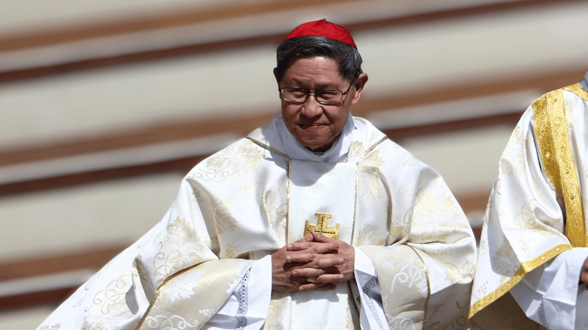 Cardeal Luis Antonio Tagle, com paramentos litúrgicos brancos e solidéu vermelho, mantém as mãos unidas durante celebração.
