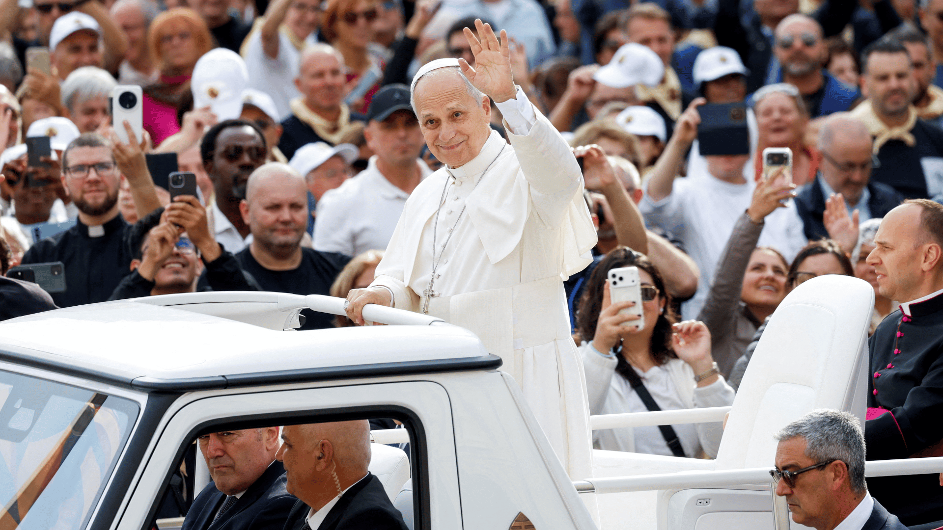 A imagem mostra o PApa Leão XIV com suas habituais vestes brancas. Ele está em pé no papamóvel e acena aos fiéis que estão ao redor na Praça São Pedro