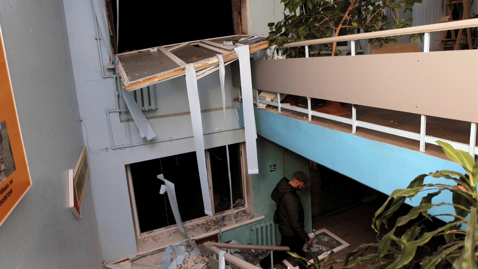 A imagem mostra um cenário de destruição em umdormitório estudantil. Há janelas quebradas e estilhaços. Também há um homem no local próximo a uma escada.