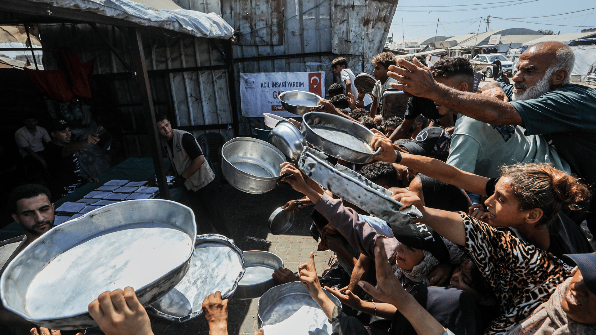 Palestinos, entre adultos e crianças, estendem recipientes em busca de alimentos durante distribuição de ajuda humanitária na Faixa de Gaza.