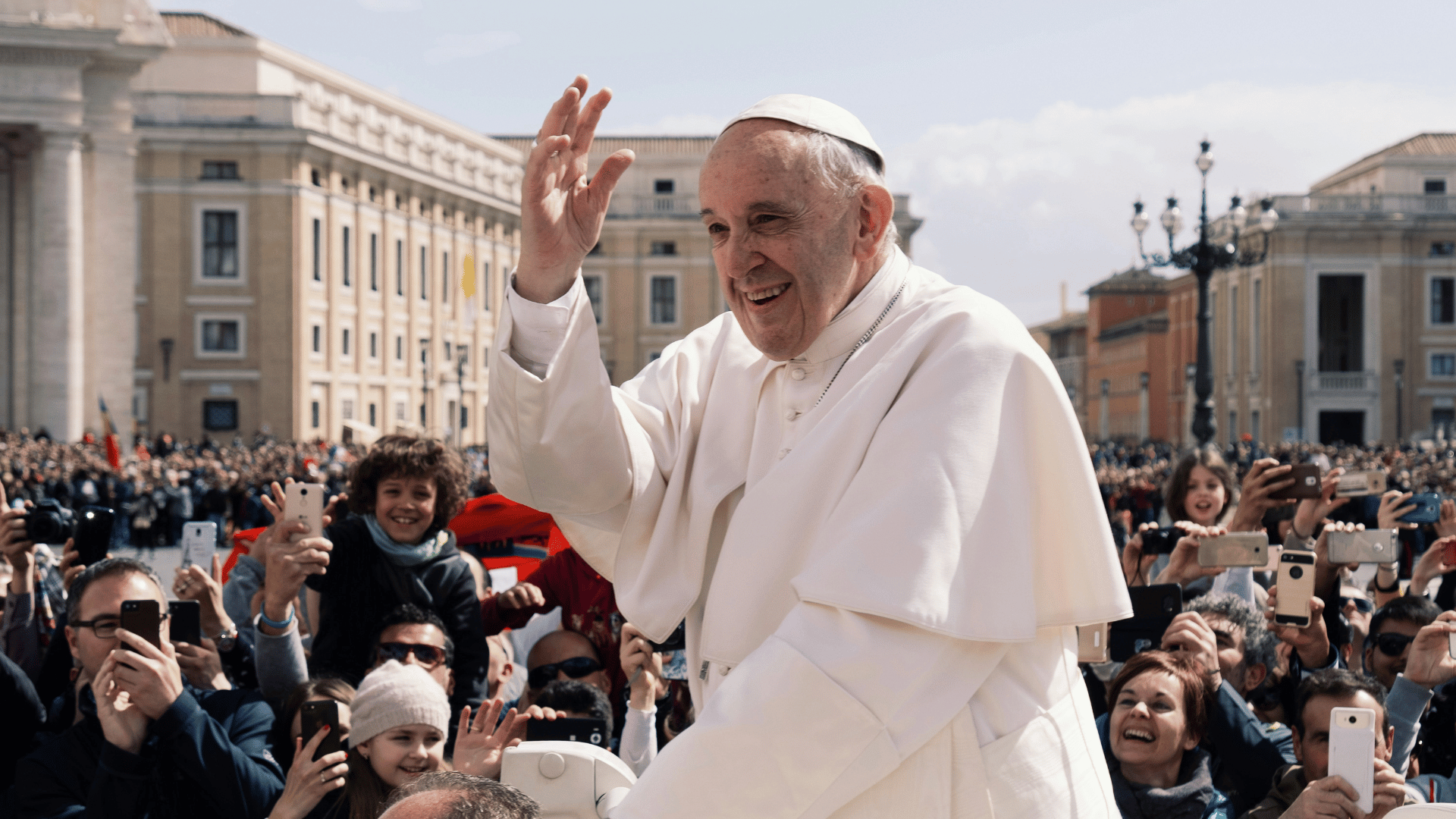 Papa Francisco, com as tradicionais vestes brancas, passa pela Praça São Pedro de papamóvel enquanto acena aos fiéis.
