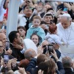 A imagem mostra o Papa Leão XIV no papamóvel em meio à multidão. Ele acaricia o rosto de um bebê erguido para que chegasse até ele.