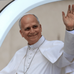 A imagem mostra o Papa Leão XIV com suas habituais vestes brancas. Ele está no papamóvel e acena aos fiéis.