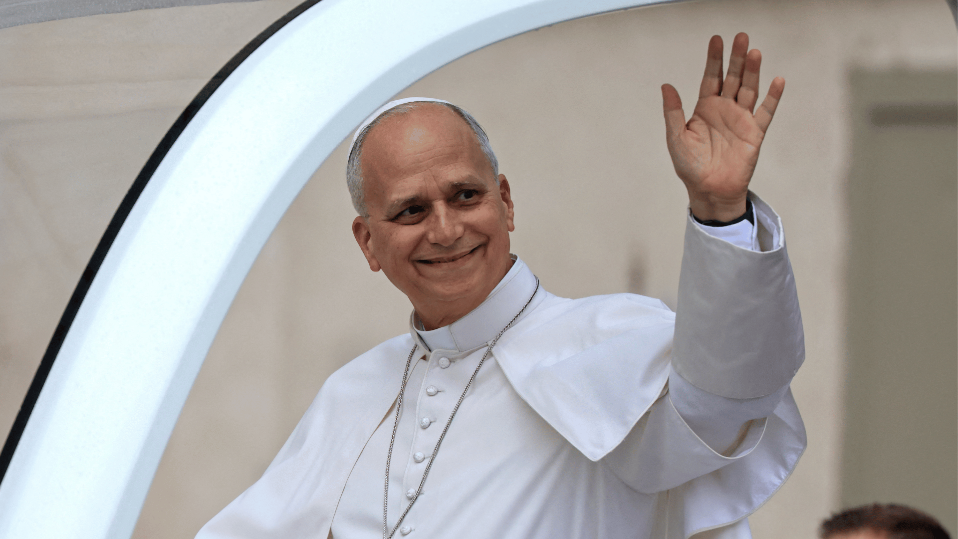 A imagem mostra o Papa Leão XIV com suas habituais vestes brancas. Ele está no papamóvel e acena aos fiéis.