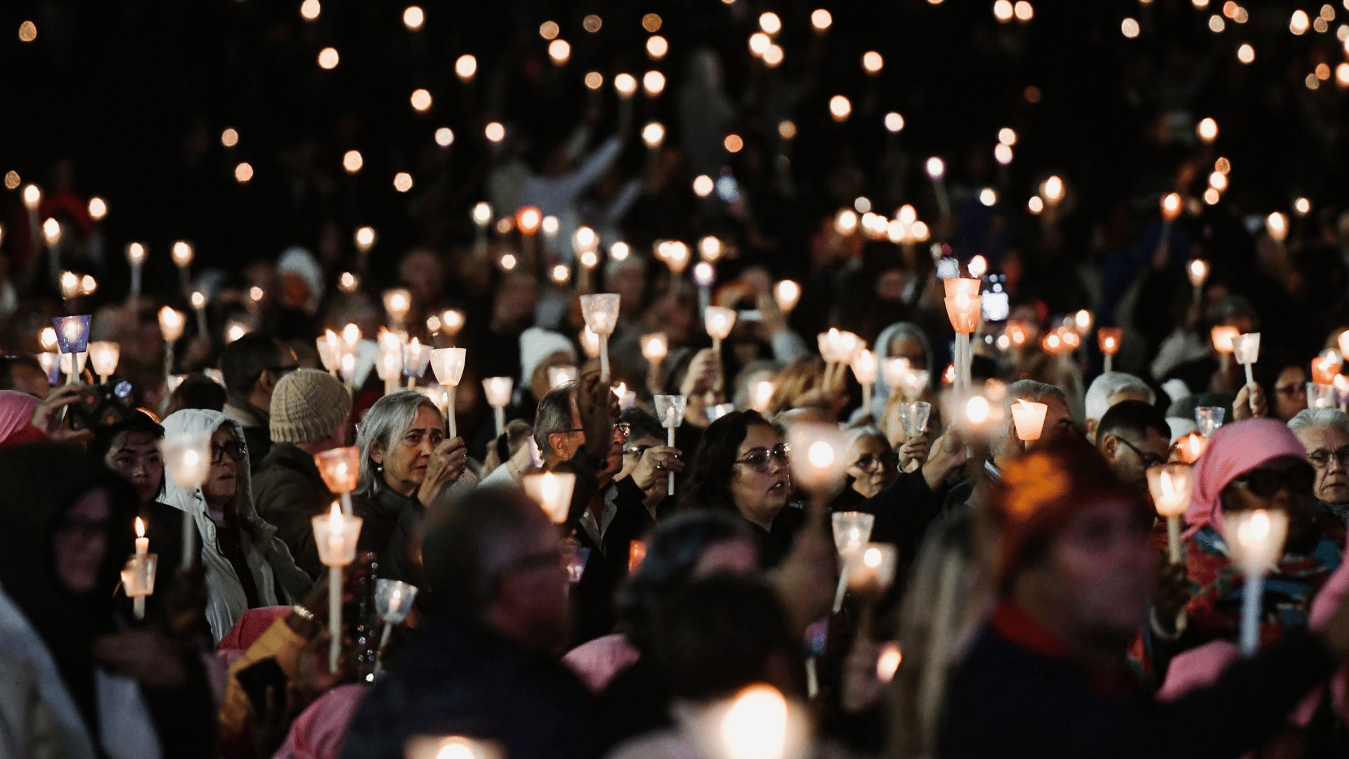 A imagem mostra uma multidão de fiéis segurando velas nas mãos. Eles estão no Santuário de Fátima em Portugal