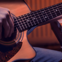 Mão tocando violão acústico em primeiro plano, com o braço do instrumento posicionado ao lado das teclas de um piano e uma folha de partitura musical sobre elas, sob iluminação acolhedora em tons quentes.