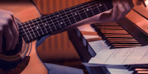 Mão tocando violão acústico em primeiro plano, com o braço do instrumento posicionado ao lado das teclas de um piano e uma folha de partitura musical sobre elas, sob iluminação acolhedora em tons quentes.