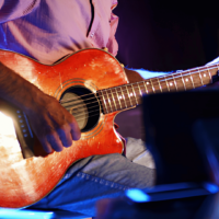 Foto em plano detalhe de um músico tocando um violão acústico de cor avermelhada e alaranjada (estilo sunburst) durante uma apresentação ao vivo em um palco. O músico, visível do torso para baixo, veste camisa listrada de manga curta e calça jeans. Sua mão esquerda segura o braço do violão, que tem um capotraste instalado, enquanto a mão direita dedilha as cordas perto do bocal. A iluminação é de show, com luzes quentes destacando o corpo do instrumento e reflexos azuis e escuros ao fundo.
