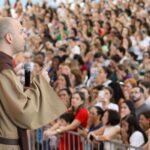 A imagem mostra Frei Gilson, um religioso, pregando para uma grande multidão. Ele está de perfil, vestido com um hábito marrom, típico dos franciscanos, e segura um microfone enquanto gesticula com a mão direita, como se estivesse enfatizando um ponto importante de sua pregação. Ao fundo, pode-se ver uma grande quantidade de pessoas, aparentemente atentas e concentradas, algumas com expressões emocionadas, outras com as mãos no rosto, indicando que estão profundamente envolvidas no que está sendo dito. As pessoas estão cercadas por uma barreira, e muitas delas seguram celulares, possivelmente registrando o momento.