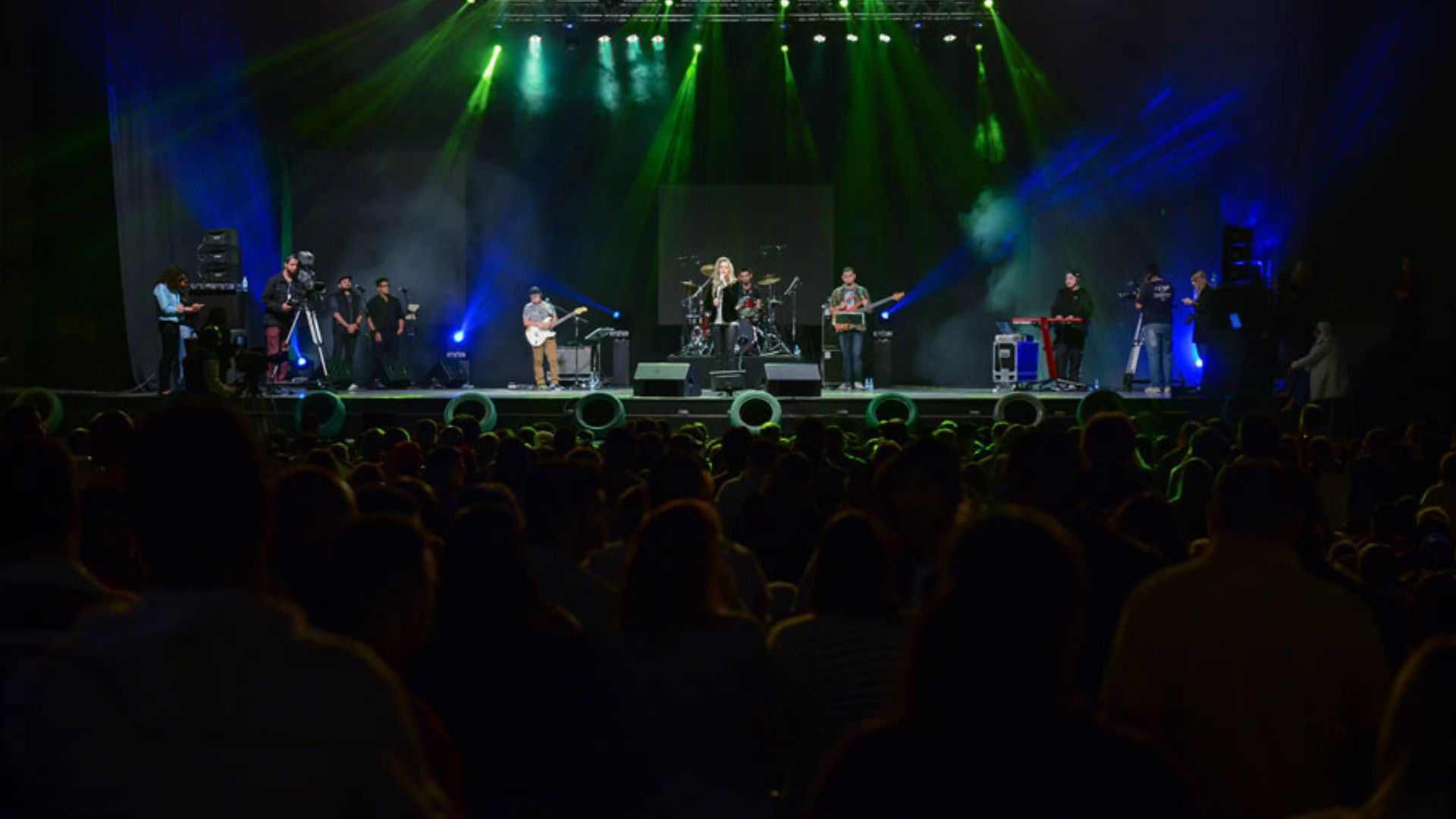 A imagem mostra uma banda tocando em um palco durante um evento musical. A cena é iluminada por luzes de palco coloridas, predominantemente verdes e azuis, que criam uma atmosfera vibrante. Há várias pessoas no palco: músicos tocando instrumentos e operadores de câmeras e equipamentos de som e luz, capturando o show. No centro do palco, uma vocalista está se apresentando, enquanto outros músicos tocam guitarra, baixo e bateria ao seu redor. O público, visto em silhueta na parte inferior da imagem, está observando o show. O ambiente parece ser um grande auditório ou arena, com uma produção técnica significativa para o evento.

