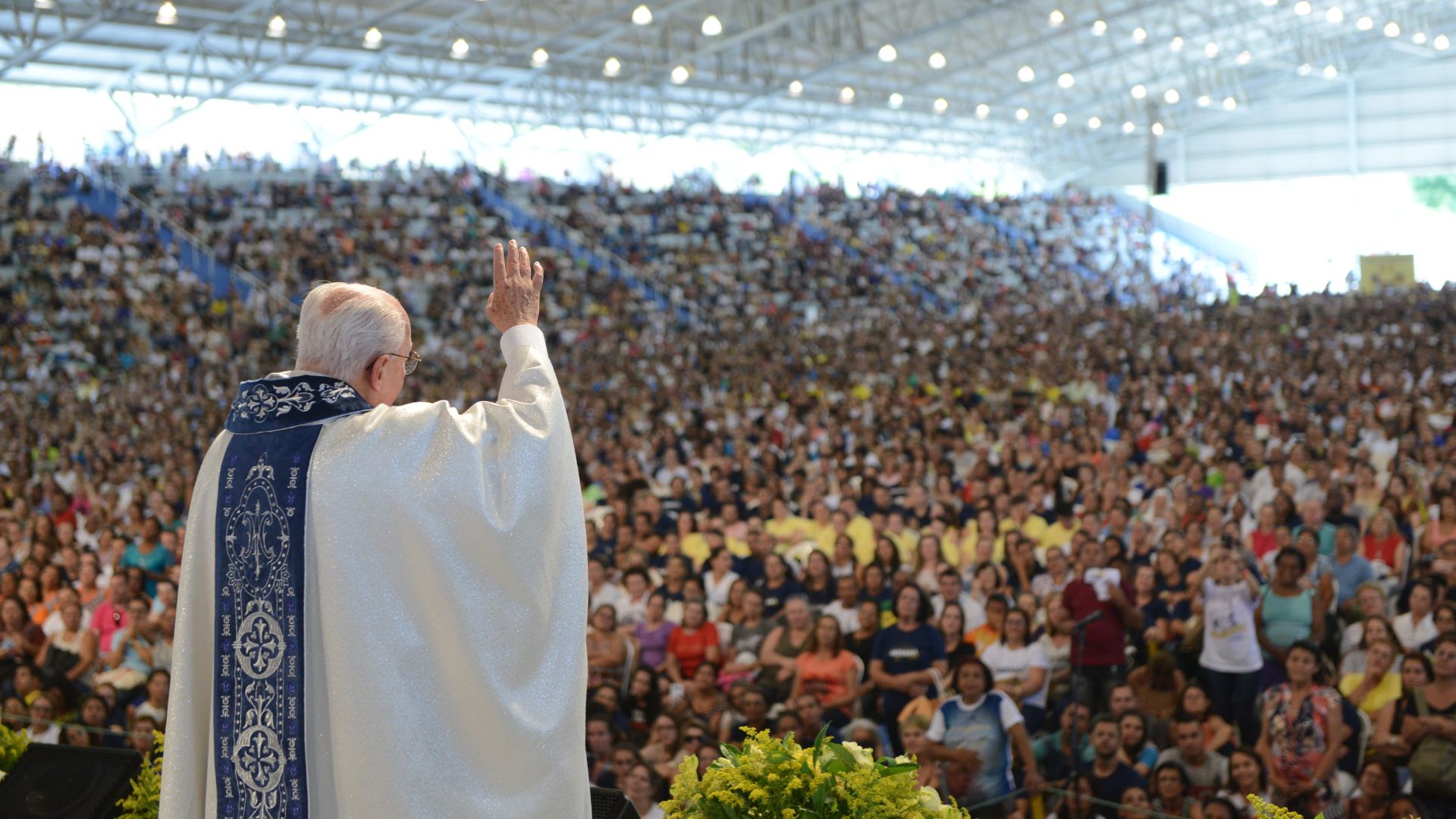 Na imagem, vemos um grande evento religioso com uma multidão reunida em um espaço coberto. Na frente da imagem, está um padre de costas para a câmera, vestido com uma batina branca e uma estola azul, com detalhes decorativos em branco. Ele está com o braço levantado em direção à multidão, como se estivesse abençoando as pessoas. A multidão, que enche o local, é composta por milhares de fiéis que parecem atentos e participativos. A estrutura do local é grande, com iluminação no teto, e a atmosfera do evento é de devoção e reverência. O padre na imagem é Monsenhor Jonas Abib, fundador da Comunidade Canção Nova.