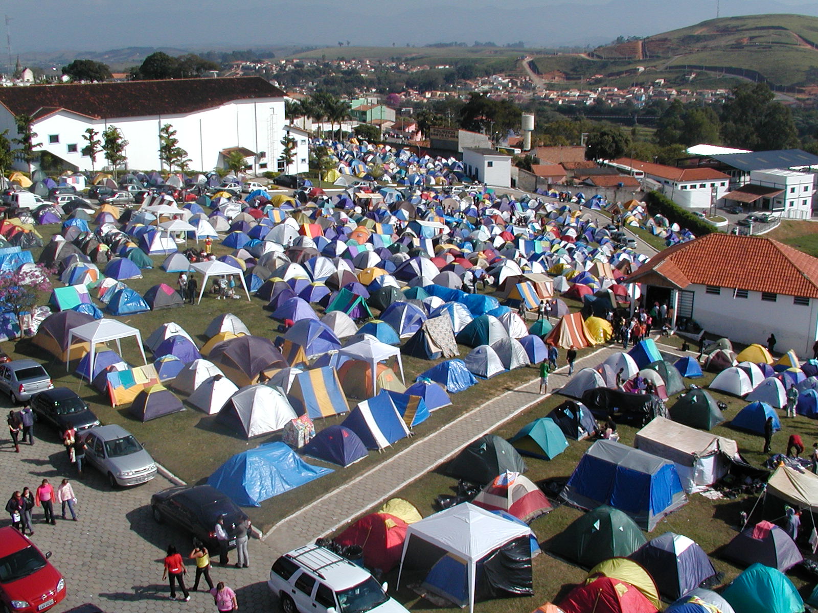 A imagem retrata o camping da Comunidade Canção Nova, em Cachoeira Paulista, SP, Brasil, durante um evento. O espaço ao ar livre está lotado de barracas coloridas de diferentes tamanhos, espalhadas por toda a área gramada e ao longo das calçadas. No fundo, é possível ver construções brancas pertencentes à comunidade, além de um estacionamento com vários carros. Ao longe, há colinas e casas, indicando a proximidade com a cidade. Muitas pessoas circulam entre as barracas, criando um ambiente movimentado e acolhedor. A Canção Nova é conhecida por acolher peregrinos em eventos religiosos, e o camping é uma forma popular de acomodação para os fiéis que participam das atividades.