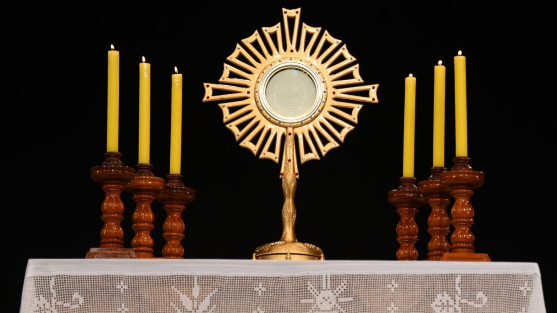 A imagem mostra um ostensório dourado em forma de sol com raios simétricos, usado para a exposição do Santíssimo Sacramento na Adoração Eucarística. O ostensório está centralizado sobre o altar, com a hóstia consagrada visível em seu centro. Ao redor, estão seis velas amarelas acesas, posicionadas em castiçais de madeira escura, três de cada lado, contribuindo para a atmosfera de reverência e solenidade típica das cerimônias de adoração. O fundo é escuro, destacando o brilho dourado do ostensório e a luz suave das velas.