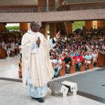 "Padre sorridente de óculos e paramentos brancos prega com microfone para uma igreja lotada de jovens em camisas azul-claras."