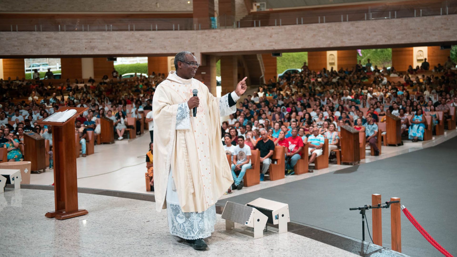 "Padre sorridente de óculos e paramentos brancos prega com microfone para uma igreja lotada de jovens em camisas azul-claras."