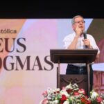 Homem idoso de camisa social branca listrada e calça jeans escura fala ao microfone em um púlpito durante evento religioso. Ao fundo, grande tela exibe o texto “A Igreja Católica e Seus Dogmas” acompanhado da imagem da cúpula da Basílica de São Pedro, no Vaticano. Na frente do púlpito, arranjo floral com rosas vermelhas e brancas.
