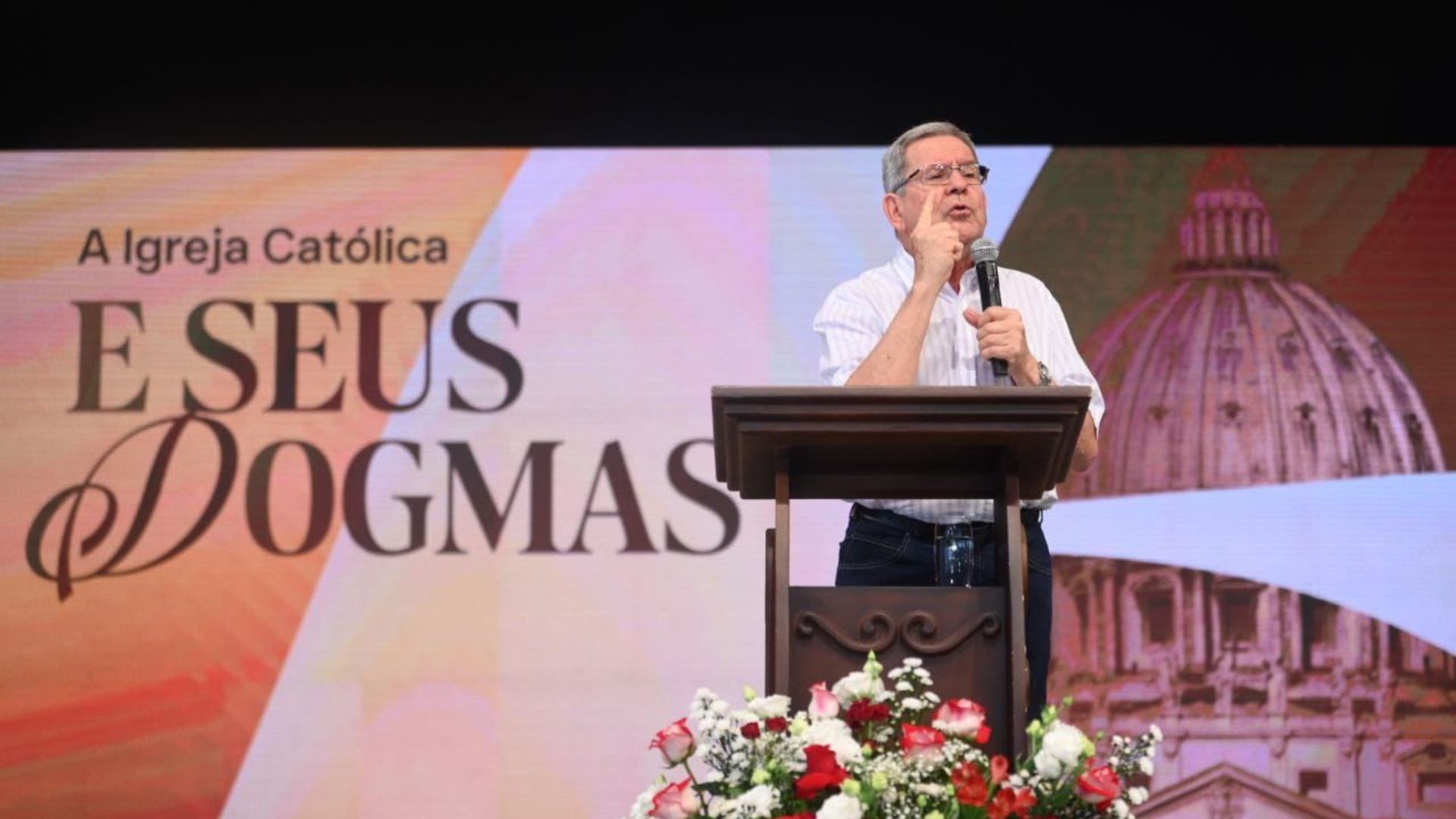 Homem idoso de camisa social branca listrada e calça jeans escura fala ao microfone em um púlpito durante evento religioso. Ao fundo, grande tela exibe o texto “A Igreja Católica e Seus Dogmas” acompanhado da imagem da cúpula da Basílica de São Pedro, no Vaticano. Na frente do púlpito, arranjo floral com rosas vermelhas e brancas.