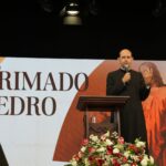 O Padre Paulo Ricardo, vestido com batina preta e clériman branco, está pregando em um púlpito de madeira durante uma palestra ou retiro. Ele segura um microfone na mão direita e faz um gesto com a mão esquerda. Ao fundo, há um grande telão com a frase “O Primado de Pedro” em letras elegantes e uma imagem clássica de Jesus entregando as chaves a São Pedro. O palco está decorado com um arranjo floral de rosas vermelhas e brancas na frente do púlpito.