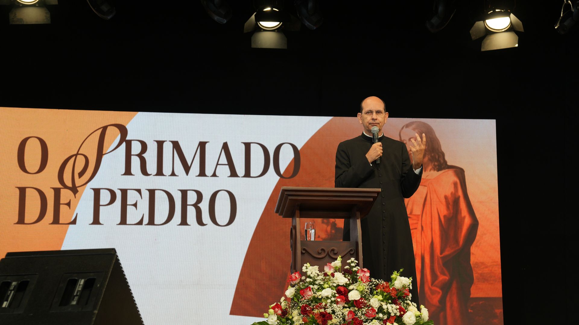 O Padre Paulo Ricardo, vestido com batina preta e clériman branco, está pregando em um púlpito de madeira durante uma palestra ou retiro. Ele segura um microfone na mão direita e faz um gesto com a mão esquerda. Ao fundo, há um grande telão com a frase “O Primado de Pedro” em letras elegantes e uma imagem clássica de Jesus entregando as chaves a São Pedro. O palco está decorado com um arranjo floral de rosas vermelhas e brancas na frente do púlpito.