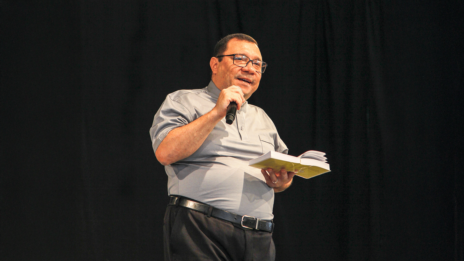 Padre Wagner Ferreira está em pé em um palco com fundo preto, iluminado por um foco de luz. Ele segura um microfone preto na mão direita, próximo à boca, e um livro aberto (aparentemente um missal ou bíblia) na mão esquerda. Usa camisa social cinza de mangas curtas dobradas, calça social escura e cinto preto. Está sorrindo levemente, com expressão serena e acolhedora, enquanto fala ou prega.
