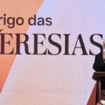 alt="Padre católico de batina preta falando ao microfone em púlpito com telão ao fundo exibindo o título 'O perigo das HERESIAS' em letras grandes sobre fundo laranja e vermelho"