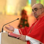 O Padre José Eduardo de Oliveira, vestindo paramentos litúrgicos vermelhos com detalhes dourados, está pregando a homilia no púlpito do Santuário Pai das Misericórdias, em Cachoeira Paulista (SP). Ele aparece de perfil, com a mão direita levantada em gesto de explicação, enquanto a esquerda repousa sobre o livro aberto. Ao fundo, é possível ver o presbitério iluminado e um árvore de Natal decorada, indicando que a celebração ocorre no tempo do Natal ou perto dessa festividade.