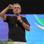 Um homem de pele clara, cabelos grisalhos curtos e óculos de armação preta está em pé no palco segurando um microfone com a mão direita e fazendo um gesto com a mão esquerda levantada, como se tocar na testa. Ele veste camiseta preta, calça clara e usa um crucifixo de madeira no pescoço. Ao fundo, há um grande telão com fundo azul e detalhes em verde e amarelo, sugerindo um evento em ambiente interno iluminado.