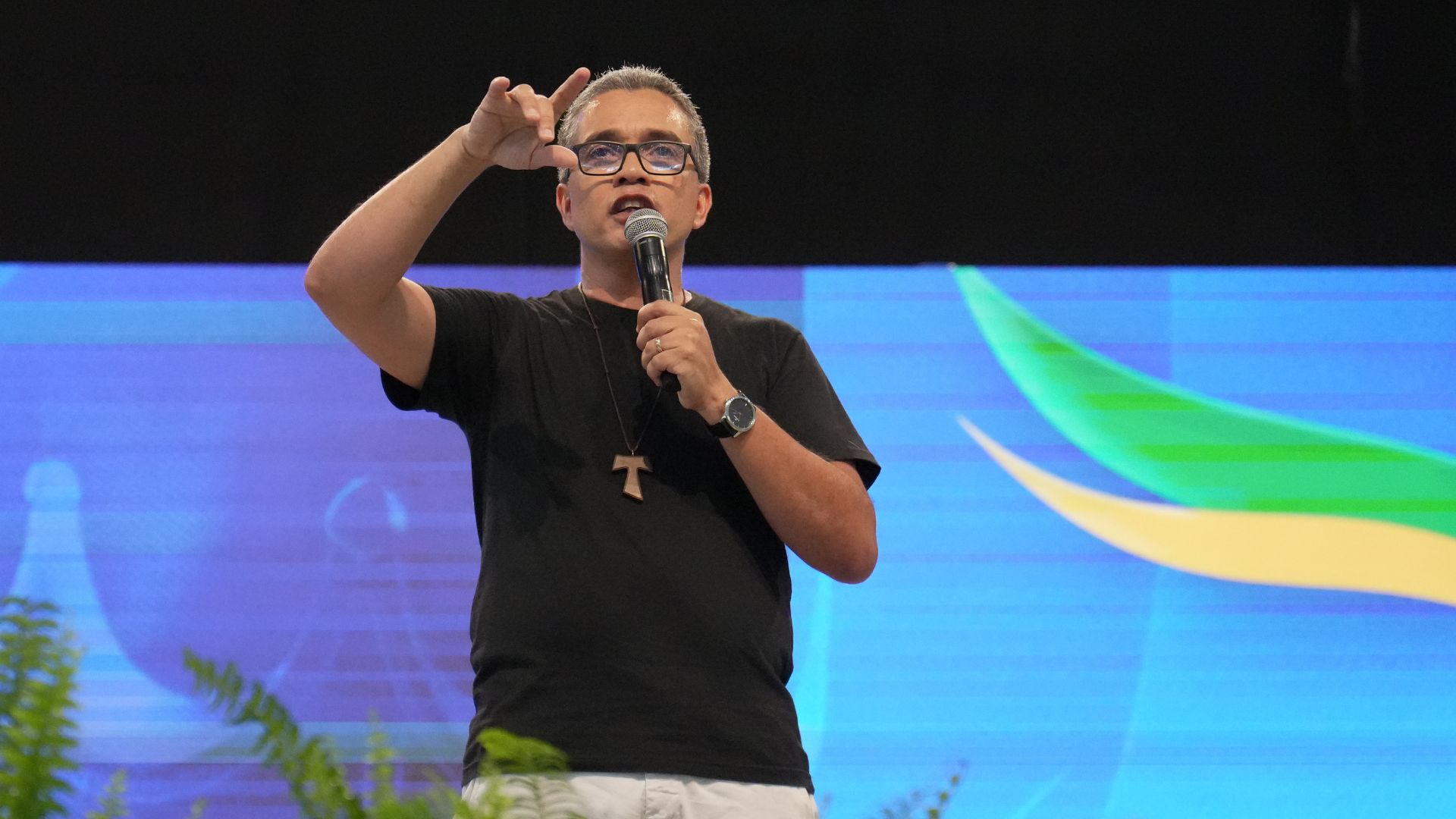 Um homem de pele clara, cabelos grisalhos curtos e óculos de armação preta está em pé no palco segurando um microfone com a mão direita e fazendo um gesto com a mão esquerda levantada, como se tocar na testa. Ele veste camiseta preta, calça clara e usa um crucifixo de madeira no pescoço. Ao fundo, há um grande telão com fundo azul e detalhes em verde e amarelo, sugerindo um evento em ambiente interno iluminado.