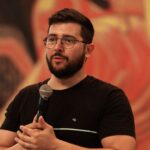 Pregador com barba e óculos, usando camiseta preta segurando um microfone na mão.