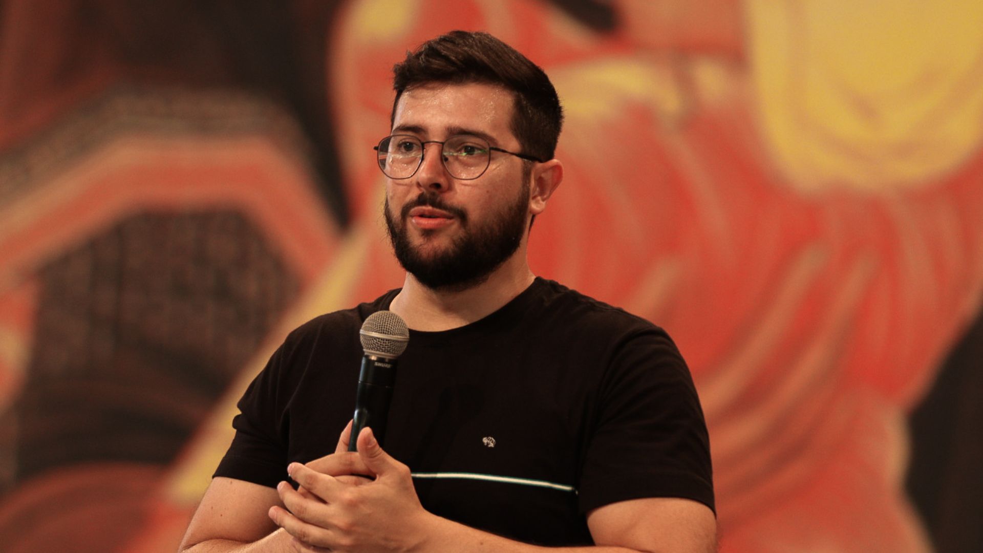 Pregador com barba e óculos, usando camiseta preta segurando um microfone na mão.