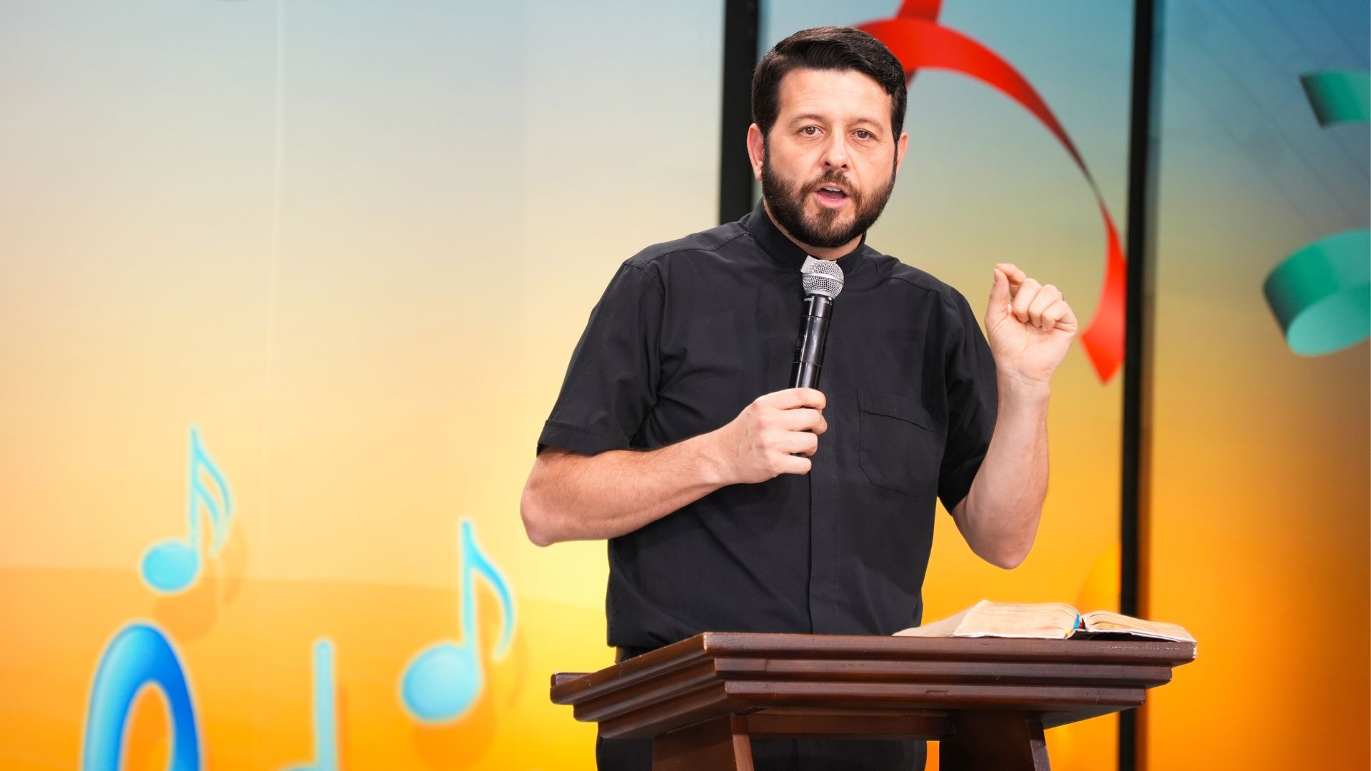 Sacerdote com camisa clerical preta segurando um microfone com a mão direita e gesticula com a mão esquerda. 
