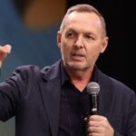 Homem com camiseta e blazer preto, com microfone na mão.