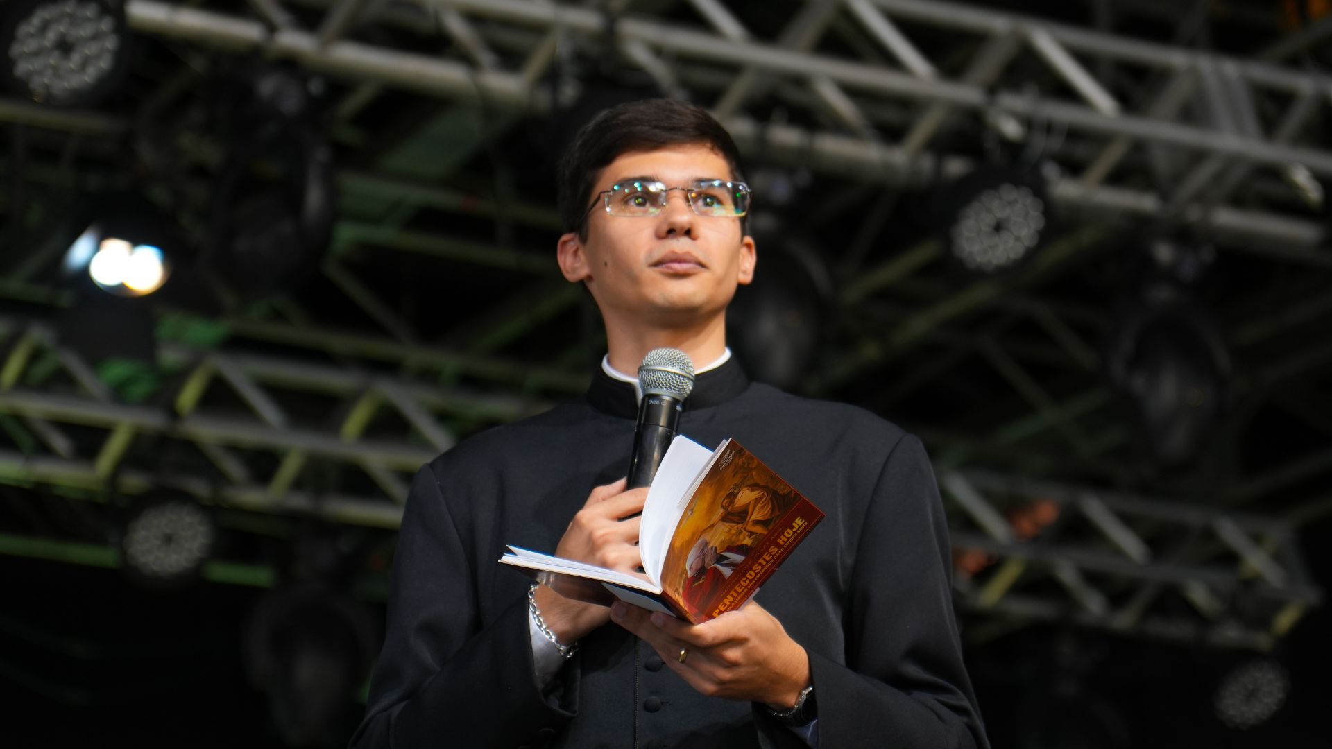 Sacerdote com batina, segurando microfone e livro nas mãos.