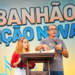 O homem, de cabelos grisalhos e óculos, fala ao microfone enquanto gesticula sobre uma Bíblia no púlpito. Ao seu lado, a jovem de blusa vermelha observa atentamente.