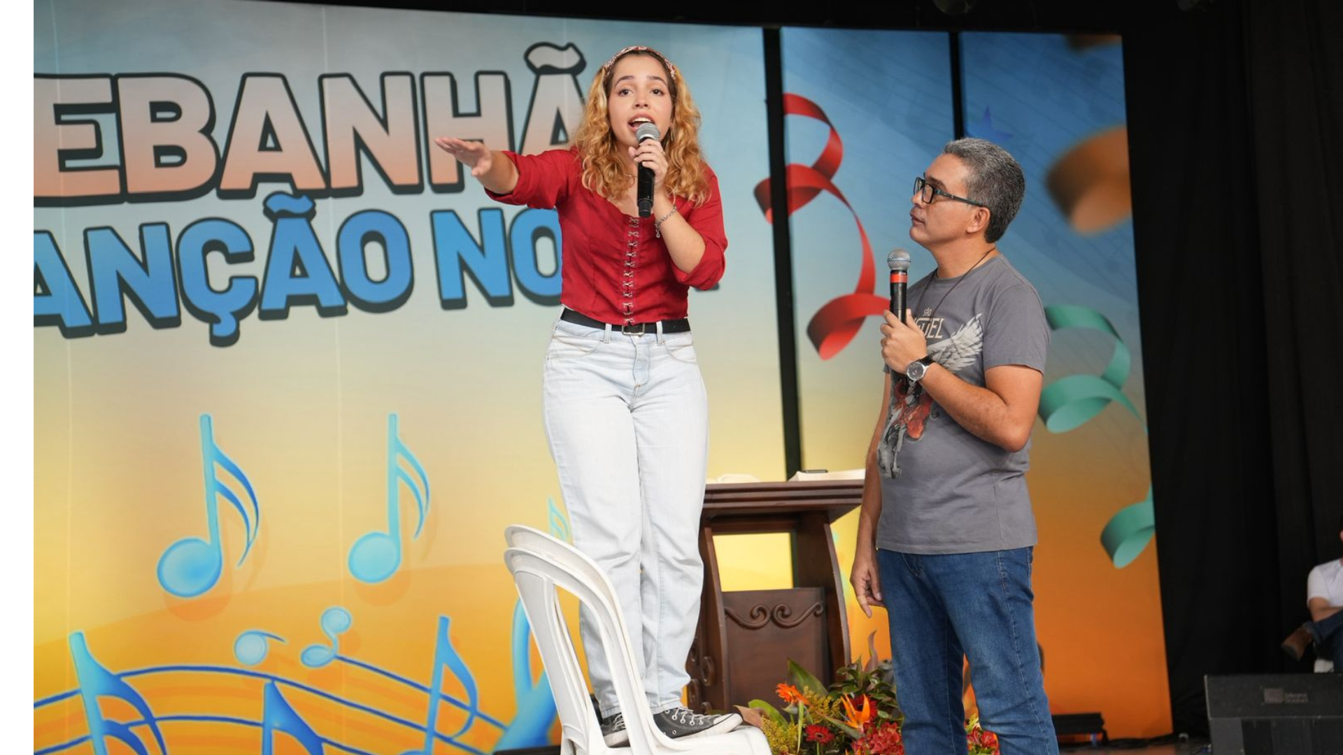 No centro, uma jovem de cabelos ondulados, vestindo uma blusa vermelha e calça clara, está em pé sobre uma cadeira de plástico branca. Ela segura um microfone e gesticula com a mão aberta. Ao lado dela, um homem de óculos e camiseta cinza observa a cena segurando outro microfone estando abaixo da jovem.