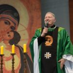 Padre com vestimenta verde com microfone na mão, ao fundo pintura de Nossa Senhora do Perpétuo Socorro.