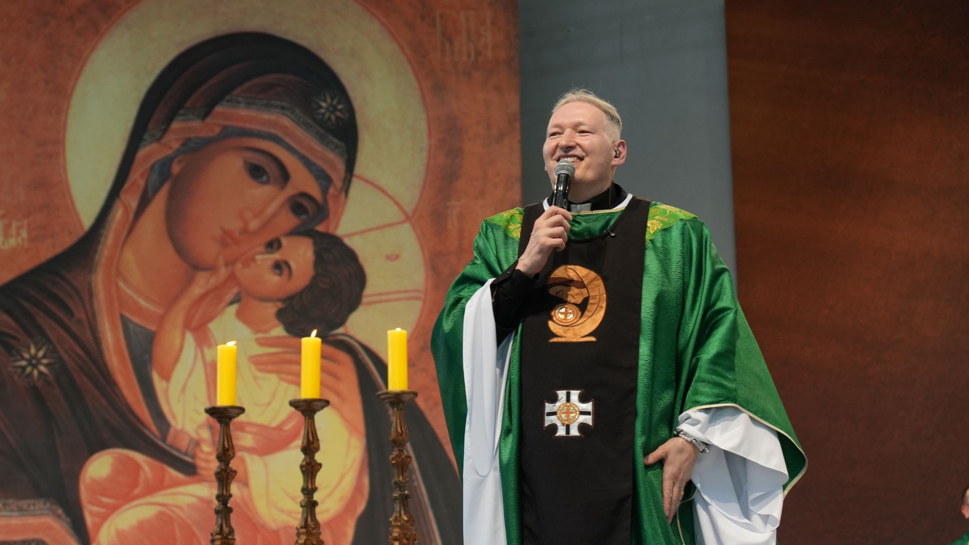 Padre com vestimenta verde com microfone na mão, ao fundo pintura de Nossa Senhora do Perpétuo Socorro.