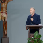 Pregador com roupa preta, segurando o microfone nas mãos atras de um púlpito.