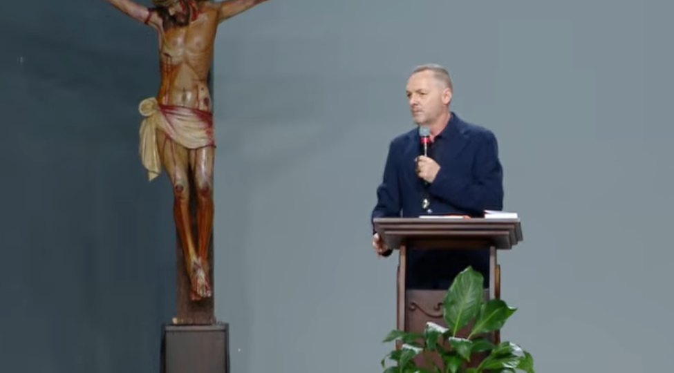 Pregador com roupa preta, segurando o microfone nas mãos atras de um púlpito. 