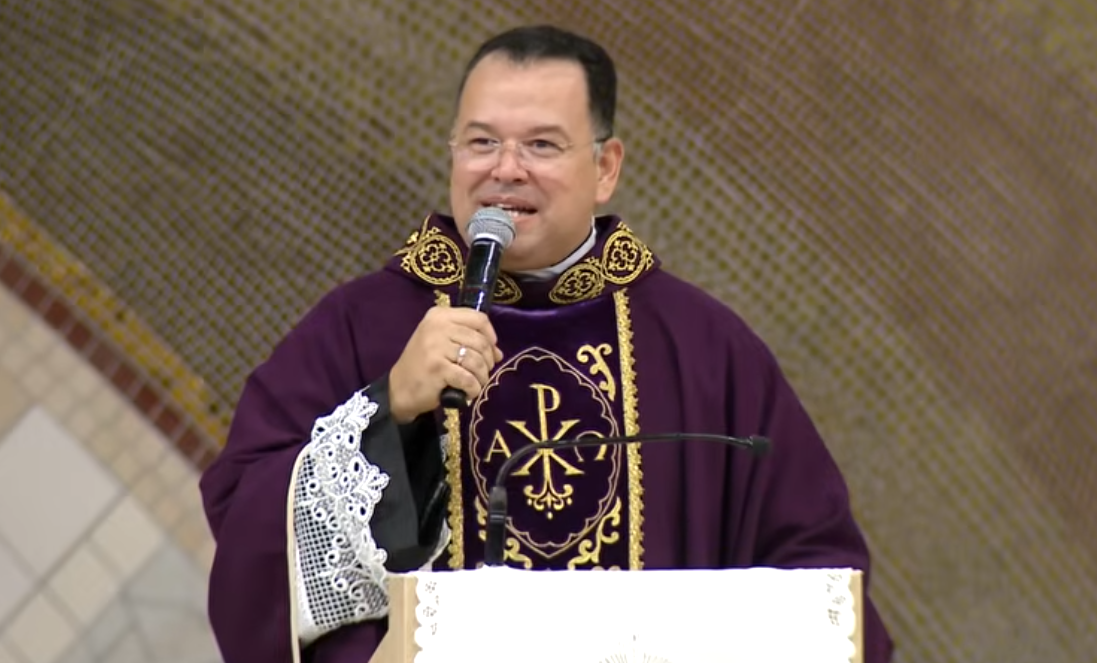 Padre com vestimenta litúrgica na cor roxa, com microfone na mão em uma igreja.