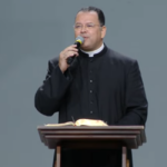 Sacerdote com microfone na mão a sua frente um púlpito e uma bíblia.