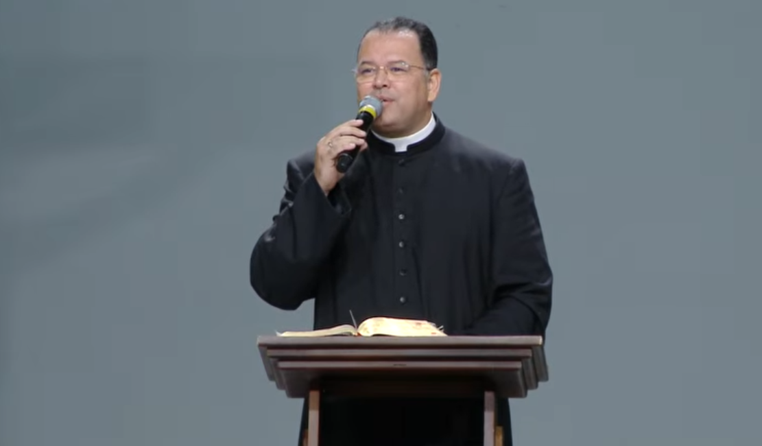 Sacerdote com microfone na mão a sua frente um púlpito e uma bíblia.