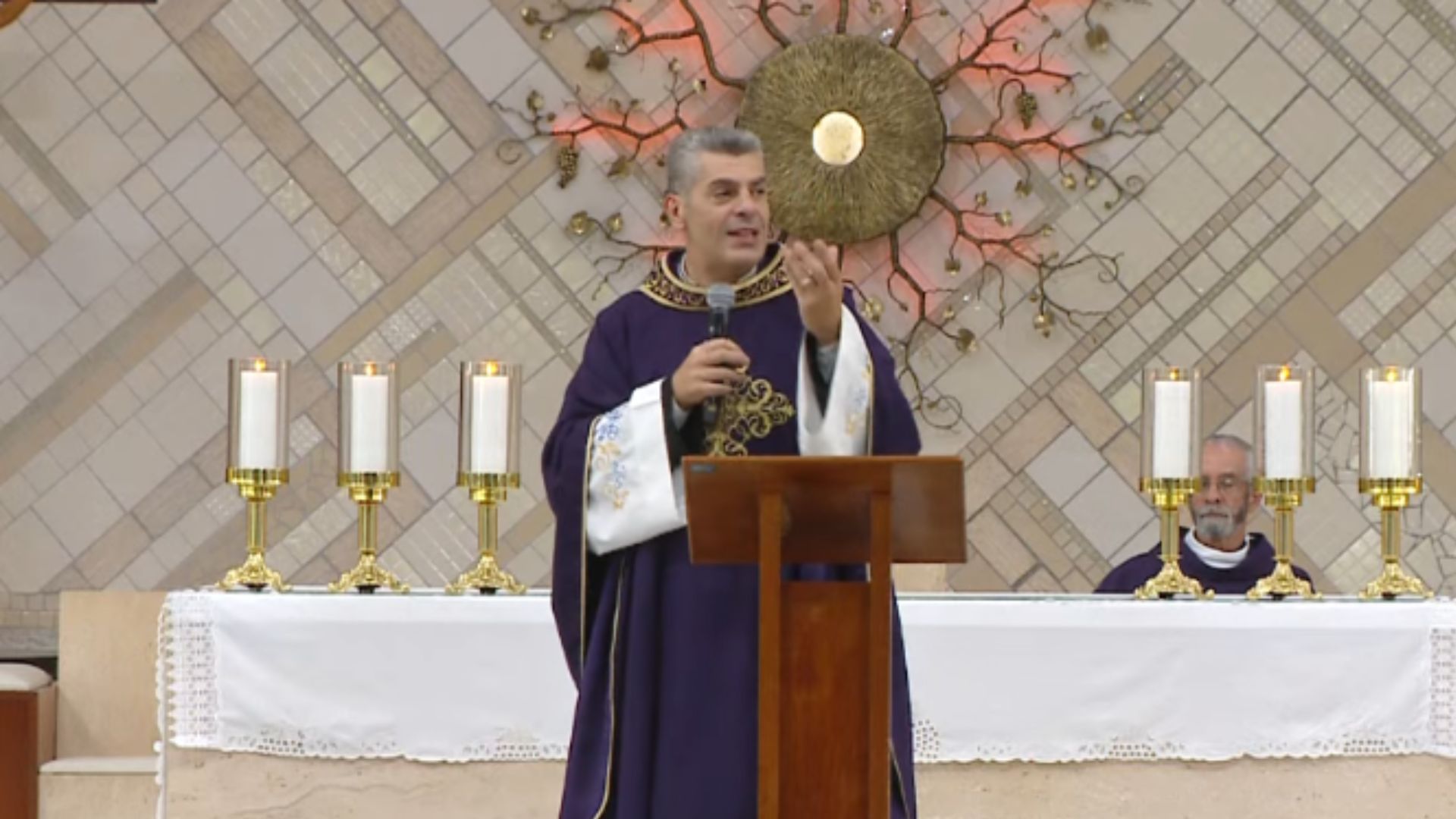 Padre com veste litúrgica roxa segurando microfone na mão.