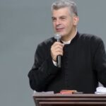 Sacerdote usando batina, atras de um púlpito segurando microfone na mão.