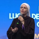 Uma mulher vestida com hábito religioso e véu branco segura um microfone enquanto fala em público. Tema: "Eucaristia, Lugar de encontro com Cristo"