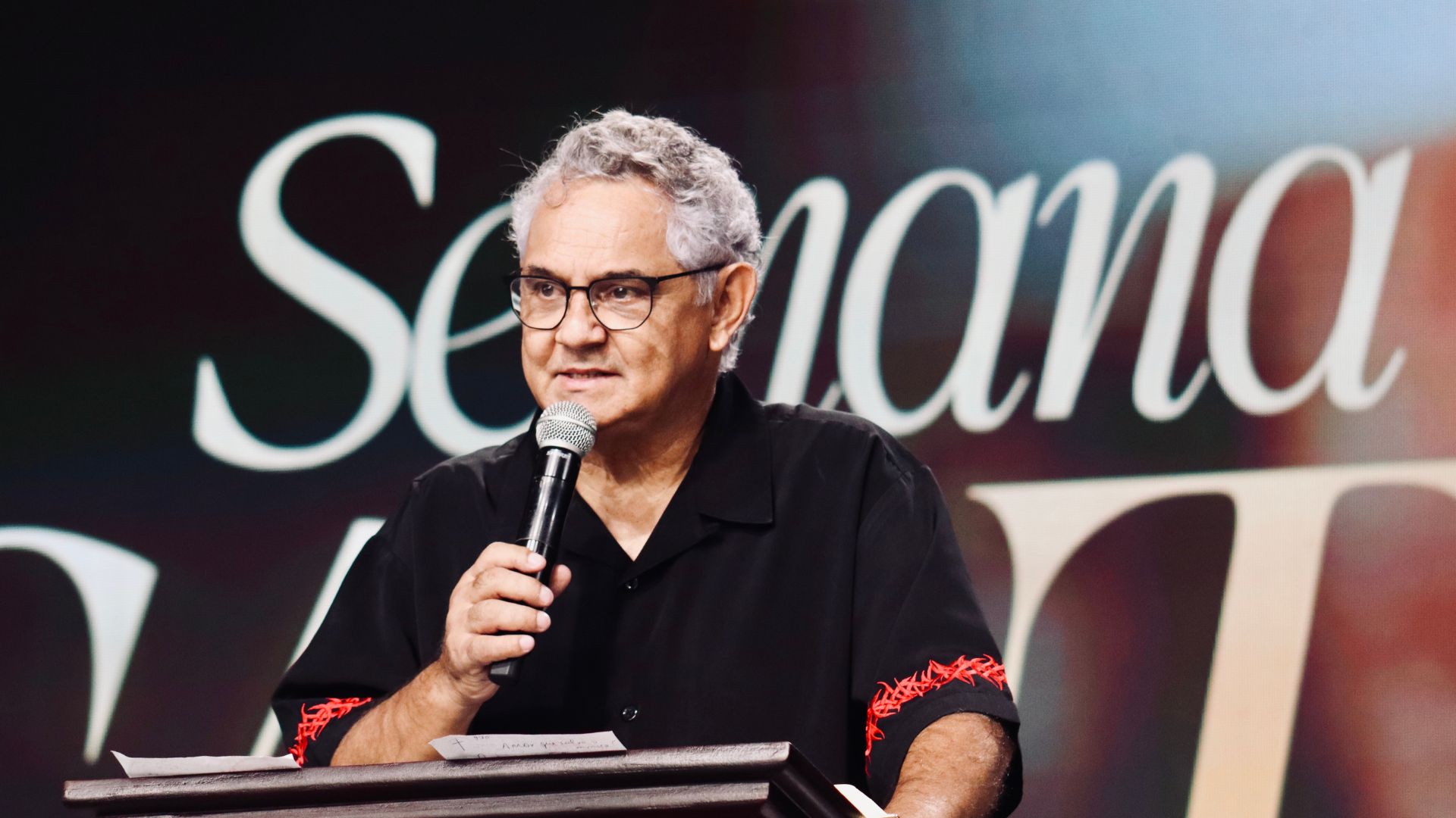Homem atrás de um púlpito em um palco, falando ao público. Ele tem cabelos grisalhos curtos, usa óculos e veste uma camisa preta com detalhes vermelhos nas mangas. Na mão direita, segura um microfone, enquanto a mão esquerda está levantada
