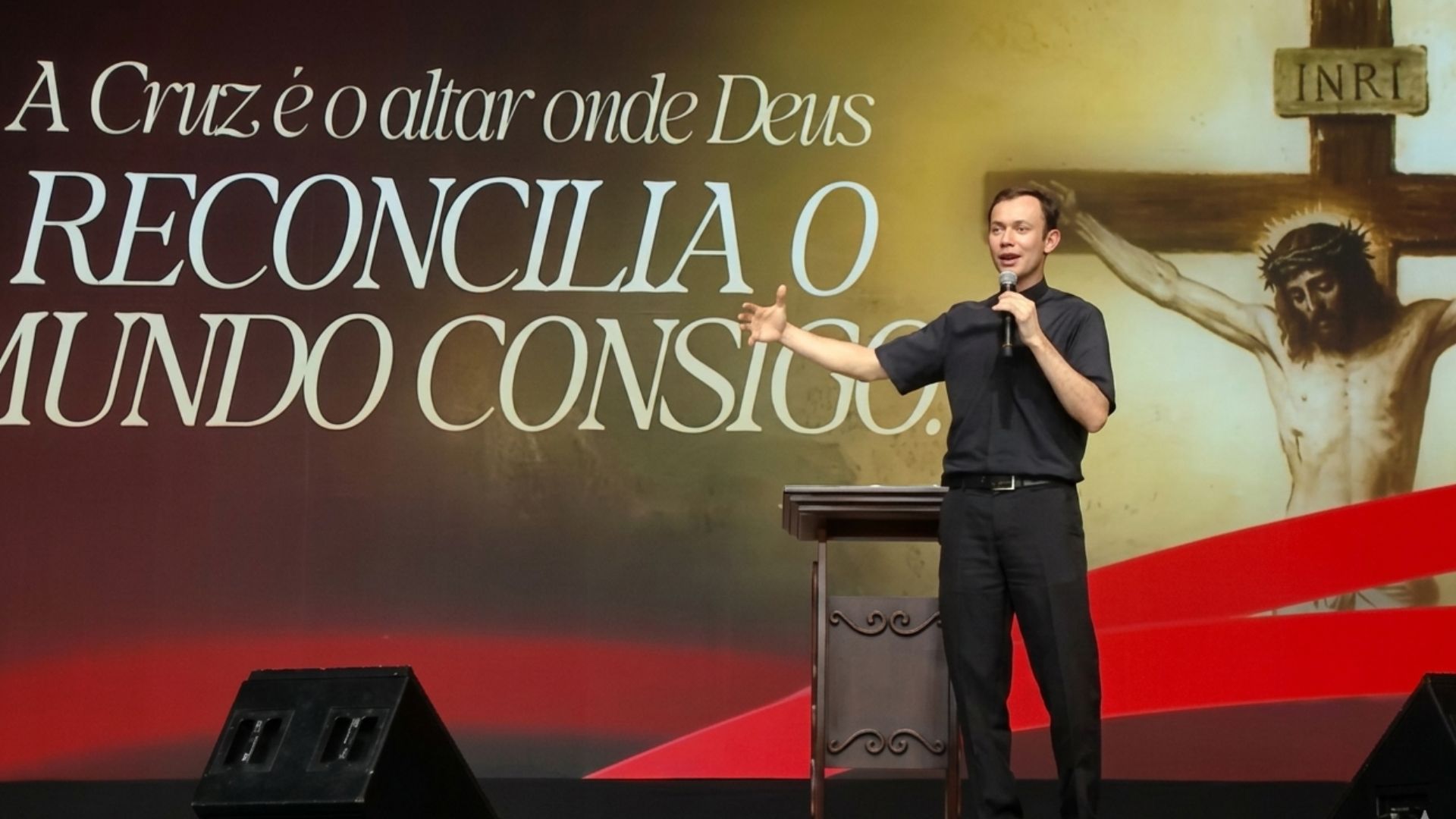 Sacerdote empé usando roupa clerica, enquanto segura com uma das mãos um microfone e gesticula com a outra, ao fundo tema da pregação escrito "A Cruz é o altar onde Deus reconcilia o mundo consigo"