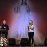 Pregador usando roupa preta segurando um crucifixo em uma das mãos e na outra o microfone atras de um púlpito. Ao lado duas mulheres, uma mulher loira de camiseta clara e calça escura cantando segurando o microfone e a outra mulher morena de blusa escura e saia estampada, cantando com as mãos levantadas. Ao fundo imagem de Jesus misericordioso representando a confiança a Jesus.