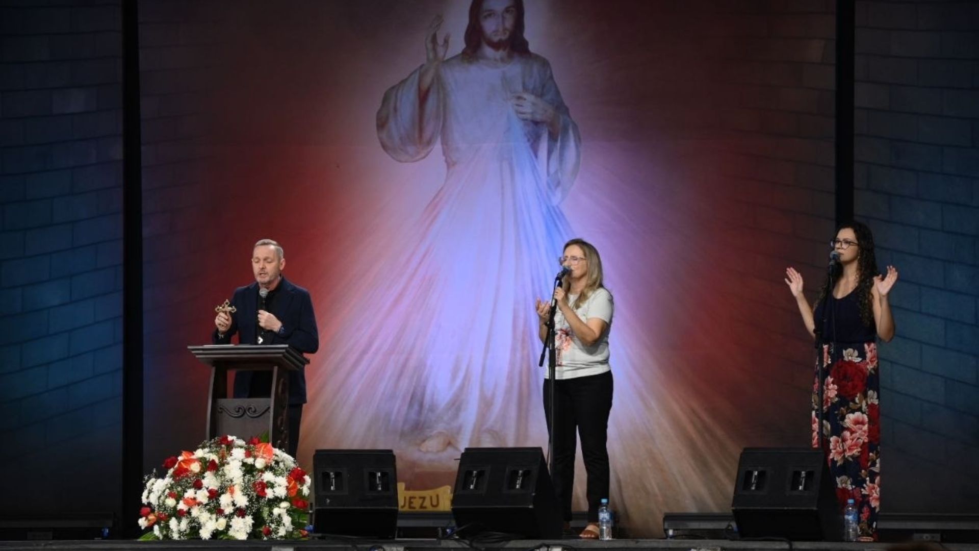 Pregador usando roupa preta segurando um crucifixo em uma das mãos e na outra o microfone atras de um púlpito. Ao lado duas mulheres, uma mulher loira de camiseta clara e calça escura cantando segurando o microfone e a outra mulher morena de blusa escura e saia estampada, cantando com as mãos levantadas. Ao fundo imagem de Jesus misericordioso representando a confiança a Jesus.