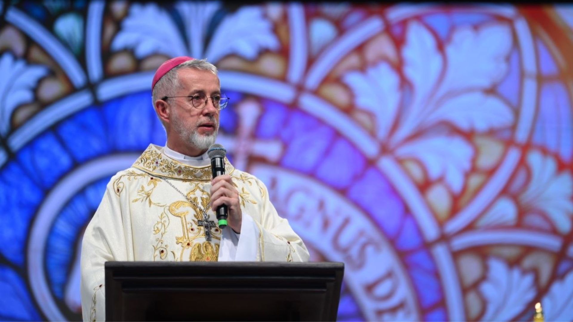 Bispo de pele clara, com barba e cabelos grisalhos, usando óculos de grau e um solidéu rosa, segurando um microfone com a mão direita, posicionado atrás de um ambão.